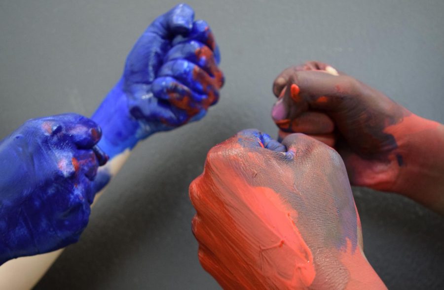Hands painted in blue and red are held in fists that have been fighting. They respectively represent opposing liberal and conservative viewpoints to showcase political polarization that stems from two sides that refuse to work to understand one another.